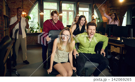 Cheerful coworkers celebrating during playful office chair race in creative workspace 142069265