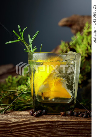 Gin and tonic cocktail with ice, lemon, juniper berries and rosemary sprigs sits on an old weathered board. Gin and tonic cocktail with ice, lemon, juniper berries and rosemary sprigs sits on an old weathered board. 142069325