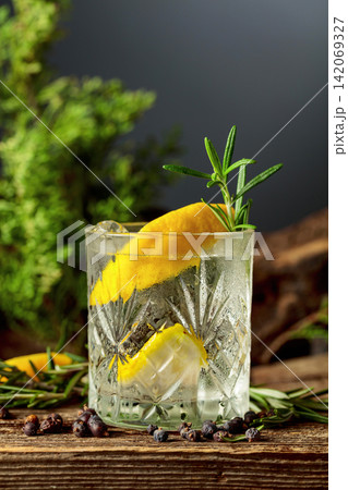 Gin and tonic cocktail with ice, lemon, juniper berries and rosemary sprigs sits on an old weathered board. 142069327