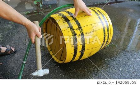 Men's hands repair or strengthen the hoops on a yellow wooden barrel with a hammer. The process takes place on wet asphalt outdoors, with a green garden hose nearby. 142069359