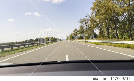 POV from moving car at sunny summer day. Through the windshield white lane marking runs steadily along asphalt road, creating a clear sense of speed, rhythm and open daytime travel POV from moving car at sunny summer day. Through the windshield white lane marking runs steadily along asphalt road, creating a clear sense of speed, rhythm and open daytime travel 142069738