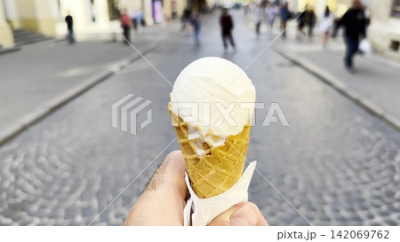 POV of male hand holding an ice cream cone while walking along city street. Blurred pedestrians pass by in the background, creating a lively urban atmosphere and emphasizing the treat in arm 142069762
