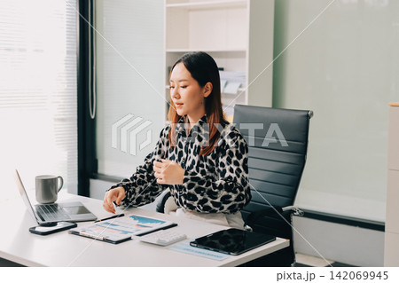 Office Discomfort: A female office worker in a red shirt experiences neck pain while working on her laptop, surrounded by office supplies and a calculator, indicating a stressful work environment. 142069945