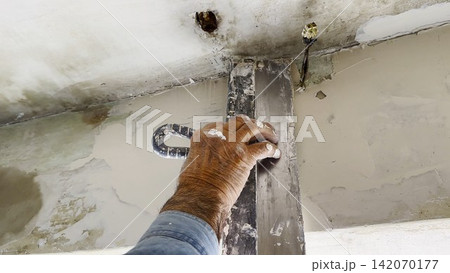 Male hand uses a putty knife to apply and spread fresh putty on interior wall. Worker carefully levels the material preparing surface for finishing and creating smooth base for further renovation work Male hand uses a putty knife to apply and spread fresh putty on interior wall. Worker carefully levels the material preparing surface for finishing and creating smooth base for further renovation work 142070177