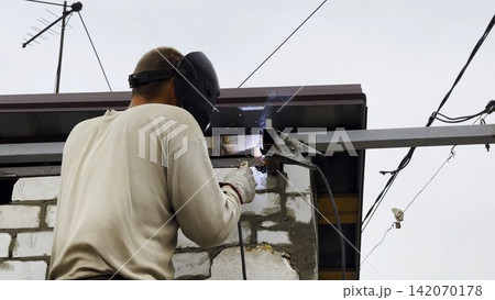 Manual laborer in protective mask welds iron detail at construction. Professional young repairman with welding machine working at the site. Male welder operating with metal on house. Close up Manual laborer in protective mask welds iron detail at construction. Professional young repairman with welding machine working at the site. Male welder operating with metal on house. Close up 142070178