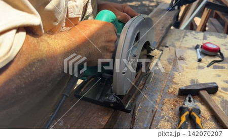 Arms of professional repairman sawing wooden construction with a circular saw at construction. Male hands of adult craftsman cuts wood using electric grind wheel at the site. Slow motion 142070207