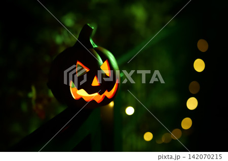 Glowing Halloween pumpkin lantern on fence at night with festive lights in background. Glowing Halloween pumpkin lantern on fence at night with festive lights in background. 142070215