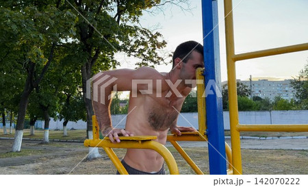 Strong sporty man exercising at sports ground. Male shirtless athlete doing push ups on parallel bars at sunny day. Young sportsman training hard outdoor. Concept of active sportive lifestyle Strong sporty man exercising at sports ground. Male shirtless athlete doing push ups on parallel bars at sunny day. Young sportsman training hard outdoor. Concept of active sportive lifestyle 142070222