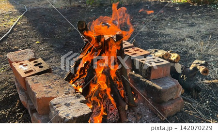 Small campfire with wooden sticks brightly burn at sunny summer day. Gentle flames and glowing embers create warm vibrant atmosphere. The scene conveys outdoor adventure and elemental beauty of fire Small campfire with wooden sticks brightly burn at sunny summer day. Gentle flames and glowing embers create warm vibrant atmosphere. The scene conveys outdoor adventure and elemental beauty of fire 142070229