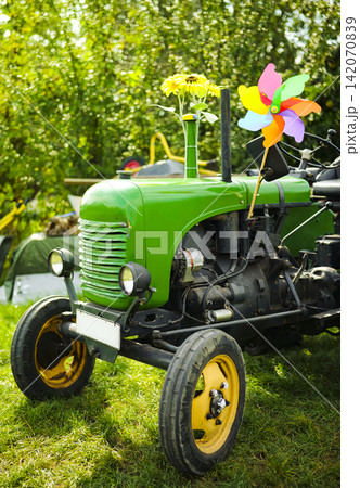 Vintage green tractor adorned with sunflowers and a pinwheel in a grassy field illustrates sustainable farming in a rural region. Vintage green tractor adorned with sunflowers and a pinwheel in a grassy field illustrates sustainable farming in a rural region. 142070839