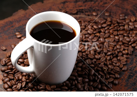 black coffee in white mug with surrounded by roasted coffee beans on dark red wood table 142071575