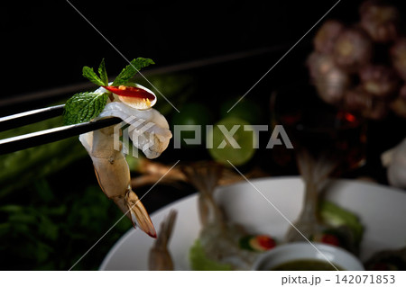 chopsticks tong a fresh raw prawn chilli and basil with another hand pouring spicy seafood sauce with a dish and seasonal in background chopsticks tong a fresh raw prawn chilli and basil with another hand pouring spicy seafood sauce with a dish and seasonal in background 142071853