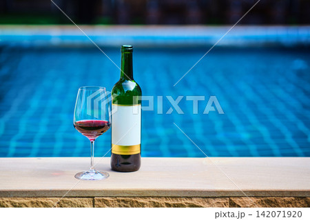 A bottle of red wine and a glass of wine are set on the edge of a clear blue pool, surrounded by a lush green lawn. The afternoon sun shines down, creating a relaxing and inviting atmosphere. 142071920