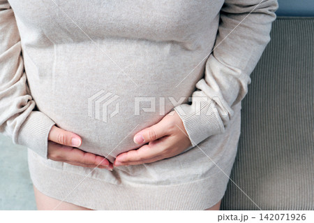 The elderly Asian woman with a pregnant belly sat on the gray sofa, gently cradling her growing baby inside. 142071926