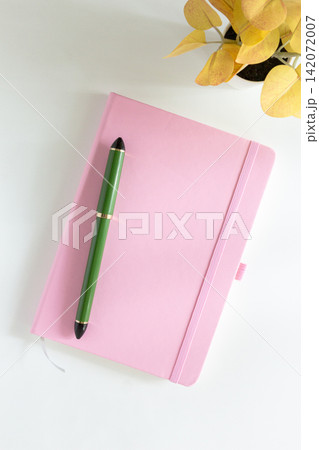 Pink notebook cover with green pen and yellow leaves plant on white desk 142072007