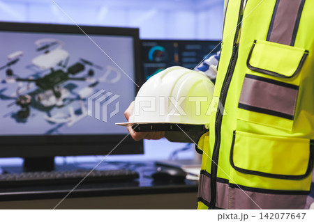 Worker in a safety vest holding a helmet beside a computer screen a drone model in an office setting Worker in a safety vest holding a helmet beside a computer screen a drone model in an office setting 142077647