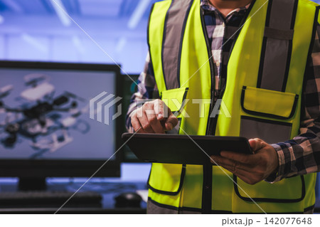 Technician in a safety vest using a tablet in a workshop with a computer monitor in the background Technician in a safety vest using a tablet in a workshop with a computer monitor in the background 142077648