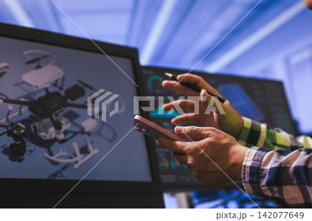 Hands using a smartphone near computer screens with a drone design on display, digital work, review, and modern technology in an office setting Hands using a smartphone near computer screens with a drone design on display, digital work, review, and modern technology in an office setting 142077649