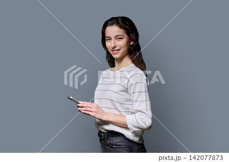 Young woman holding smartphone in half turn and looking at camera over grey background Young woman holding smartphone in half turn and looking at camera over grey background 142077873