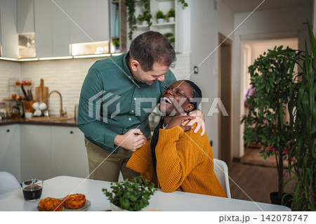 Happy diverse couple laughing and embracing at kitchen table 142078747