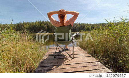 A red haired person in a pink shirt sits in a green camping chair on a wooden dock, facing away with hands clasped behind their head. A red haired person in a pink shirt sits in a green camping chair on a wooden dock, facing away with hands clasped behind their head. 142079457