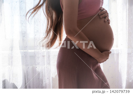 Beautiful maternity photography showing a pregnant woman gently touching her baby bump at home. Warm natural light and peaceful atmosphere representing pregnancy, motherhood, and expecting a baby. 142079739