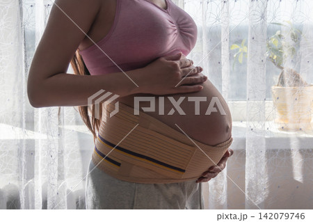 Close-up maternity photo of a pregnant woman wearing a supportive belly band and gently holding her baby bump. Concept of pregnancy care, comfort, health, and expecting motherhood. Close-up maternity photo of a pregnant woman wearing a supportive belly band and gently holding her baby bump. Concept of pregnancy care, comfort, health, and expecting motherhood. 142079746