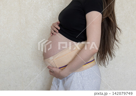 Close-up maternity photo of a pregnant woman wearing a supportive belly band and gently holding her baby bump. Concept of pregnancy care, comfort, health, and expecting motherhood. 142079749