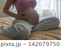 Close-up maternity portrait of a pregnant woman relaxing on a bed and touching her baby bump. Warm home atmosphere representing pregnancy, comfort, motherhood, and expecting a baby. 142079750
