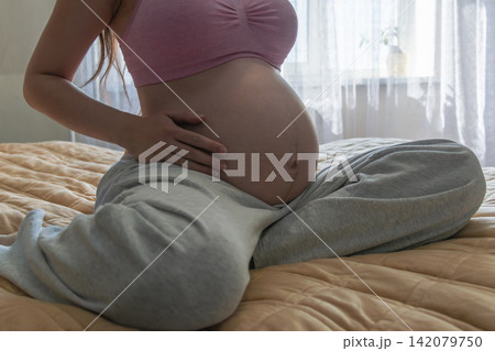 Close-up maternity portrait of a pregnant woman relaxing on a bed and touching her baby bump. Warm home atmosphere representing pregnancy, comfort, motherhood, and expecting a baby. 142079750