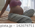 Close-up maternity portrait of a pregnant woman relaxing on a bed and touching her baby bump. Warm home atmosphere representing pregnancy, comfort, motherhood, and expecting a baby. 142079751