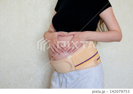 Pregnant woman showing love and connection with her unborn baby by making a heart shape with her hands on her belly. Maternity support belt, prenatal care, pregnancy health and motherhood concept. 142079753
