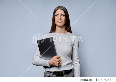 Portrait of serious young woman holding closed laptop computer on grey background Portrait of serious young woman holding closed laptop computer on grey background 142080211