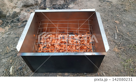 Close up of a two toned metal smoker box on dirt ground, filled with reddish wood chips beneath a metal grid perfect for outdoor smoking and campfire cooking. Close up of a two toned metal smoker box on dirt ground, filled with reddish wood chips beneath a metal grid perfect for outdoor smoking and campfire cooking. 142080231