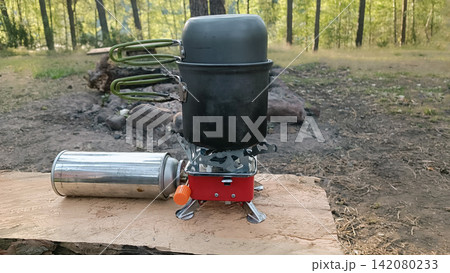 Dark gray camping pot set with olive handles cooks on a red and silver portable gas stove beside a silver fuel canister, on a wooden surface in a blurred outdoor setting. Dark gray camping pot set with olive handles cooks on a red and silver portable gas stove beside a silver fuel canister, on a wooden surface in a blurred outdoor setting. 142080233
