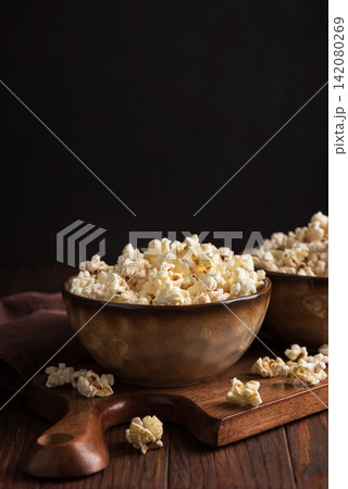 Two rustic ceramic bowls brimming with fluffy popcorn sit on a wooden cutting board with a dark background providing dramatic contrast and highlighting texture Two rustic ceramic bowls brimming with fluffy popcorn sit on a wooden cutting board with a dark background providing dramatic contrast and highlighting texture 142080269