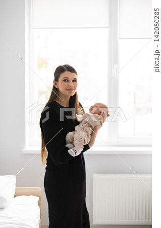 A young mother lovingly cradles her newborn baby in her arms, standing near a window with soft natural light illuminating the scene 142080285