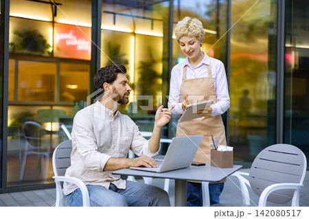 A man with a beard sits at a table with a laptop while a waitress in an apron holds a tablet and takes his order outside a cafe A man with a beard sits at a table with a laptop while a waitress in an apron holds a tablet and takes his order outside a cafe 142080517