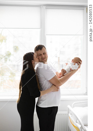 A loving mother kisses her husband while he holds their newborn baby in his arms near a window 142080533