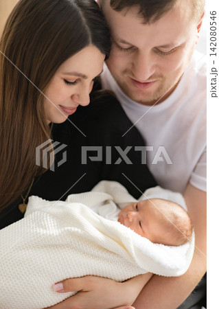 A loving mother and father embrace their newborn baby wrapped in a soft white blanket, sharing a tender moment of family connection A loving mother and father embrace their newborn baby wrapped in a soft white blanket, sharing a tender moment of family connection 142080546