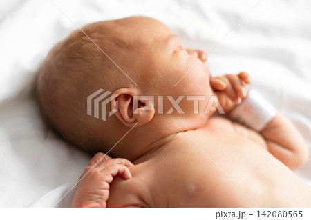 A close-up profile view of a peaceful newborn baby sleeping on a white blanket, with its hand near its mouth 142080565