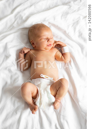 A peaceful newborn baby with closed eyes rests comfortably on a white, wrinkled sheet, wearing a diaper and hospital bracelet 142080566