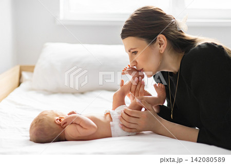 A tender moment as a mother lovingly kisses her newborn baby's tiny feet while lying on a bed A tender moment as a mother lovingly kisses her newborn baby's tiny feet while lying on a bed 142080589