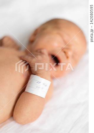 A newborn infant with a hospital identification bracelet on its arm cries while lying on a white sheet 142080595