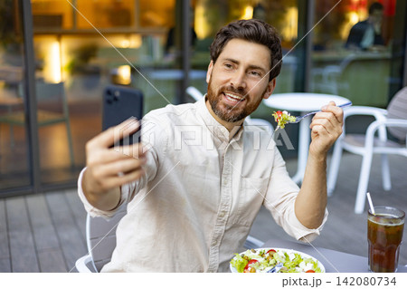 A smiling man with a beard takes a selfie with his smartphone while enjoying a fresh salad and a drink at an outdoor cafe A smiling man with a beard takes a selfie with his smartphone while enjoying a fresh salad and a drink at an outdoor cafe 142080734