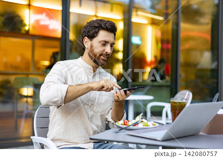 A man enjoys a meal outdoors while using his phone and working on a laptop at a cafe table A man enjoys a meal outdoors while using his phone and working on a laptop at a cafe table 142080757