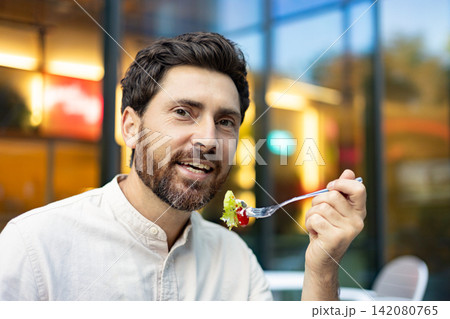 A smiling man with a beard enjoys a fresh salad with a fork while sitting outside a cafe with blurred lights in the background A smiling man with a beard enjoys a fresh salad with a fork while sitting outside a cafe with blurred lights in the background 142080765