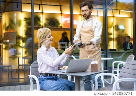 A woman uses her smartphone to make a contactless payment to a waiter at an outdoor cafe table with a laptop and salad 142080771