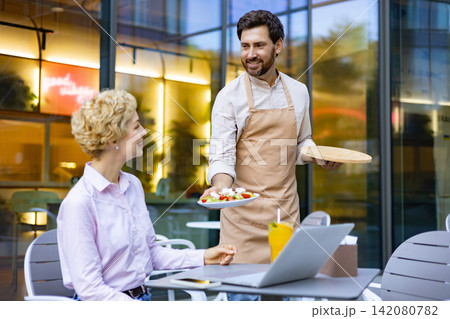 A smiling waiter in an apron presents a fresh salad to a woman seated at an outdoor cafe table with a laptop A smiling waiter in an apron presents a fresh salad to a woman seated at an outdoor cafe table with a laptop 142080782