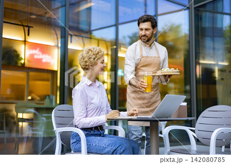 A smiling waiter presents a refreshing orange drink and a small plate of food to a woman seated at a table outside a modern cafe A smiling waiter presents a refreshing orange drink and a small plate of food to a woman seated at a table outside a modern cafe 142080785